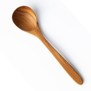 A high-angle studio shot of a handcrafted 18cm teak wood soup spoon on a clean white background. The spoon features a deep, rounded bowl and a smooth, tapered handle, showcasing the rich natural golden-brown grain and organic texture of the wood.