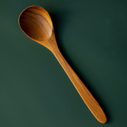 A top-down studio shot of an 18cm handcrafted teak wood soup spoon on a deep forest green background. The lighting emphasizes the deep, rounded bowl and the intentional curves of the handle, showcasing the rich, natural wood grain.