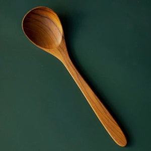 A top-down studio shot of a handcrafted 18cm teak wood soup spoon set against a deep forest green background. The lighting highlights the smooth, polished finish and the rich, swirling natural wood grain of the bowl and handle.