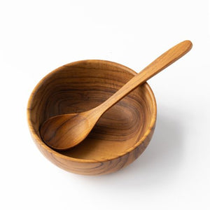 A top-down product shot of a handcrafted teak wood soup spoon resting across a matching solid teak bowl, showcasing the intricate natural wood grain and smooth, food-safe finish against a crisp white background.