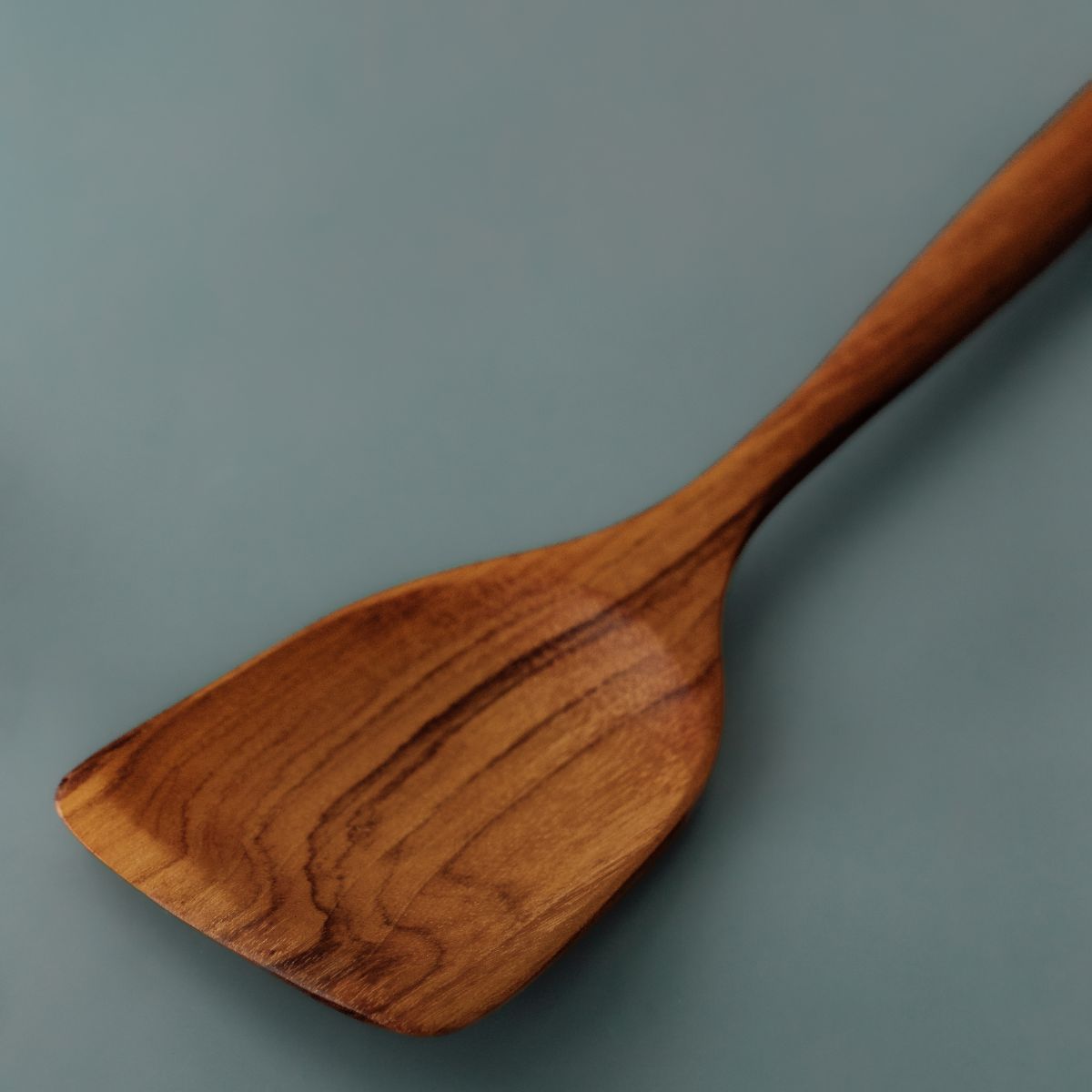 A detailed close-up shot highlighting the deep golden-brown timber grain and smooth, carved finish of a handcrafted teak spatula. Naturally water-resistant and durable, teak kitchenware maintains its rich appearance and protective surface seal when treated regularly with a food-safe beeswax conditioner.