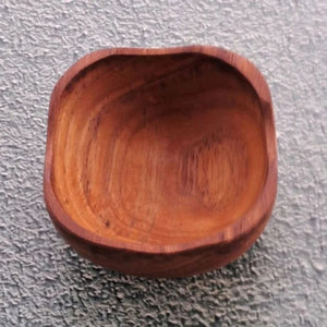 A top-down product photograph of a small, square handcrafted pinch bowl carved from sustainable teak wood, centered against a neutral, textured grey wall surface. The image highlights the unique, softly rounded square rim and deep, smooth interior of the dish, showcasing the rich golden-brown timber tones and prominent natural wood grain patterns. Expertly shaped from a single piece of non-toxic, food-safe teak, this durable mini bowl is designed as a functional culinary essential for holding spices, salts,