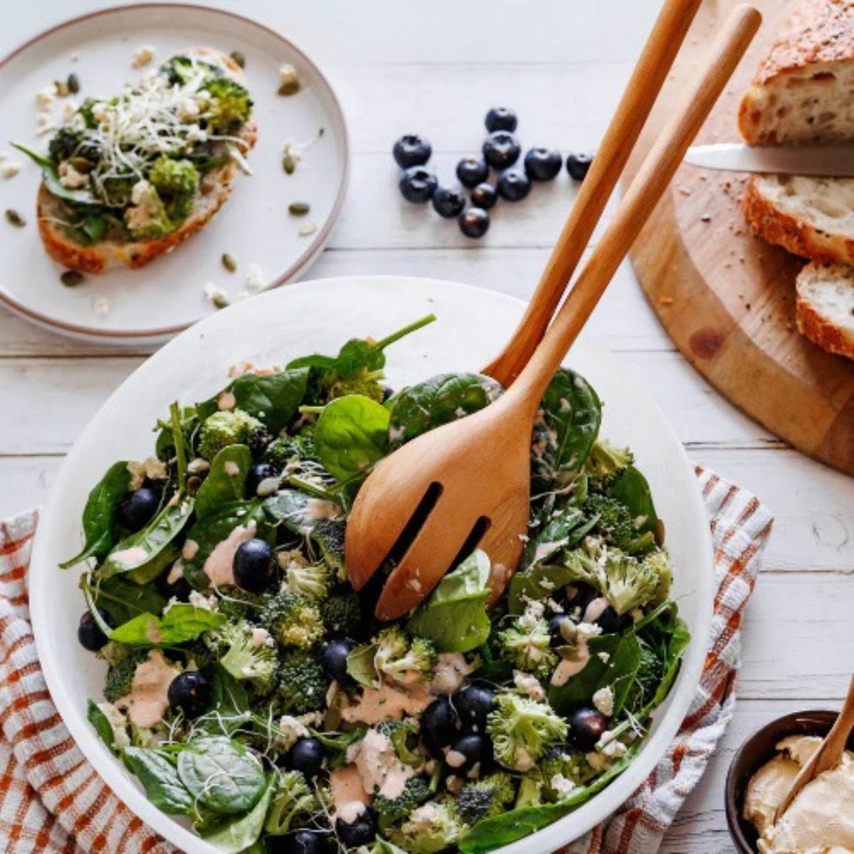 An overhead lifestyle photo of large teak wood salad servers in a white bowl of broccoli, spinach, and blueberry salad. The scene is set on a white wooden table with a side plate of avocado toast, loose blueberries, and sliced sourdough bread, showcasing a healthy, sustainable dining experience.