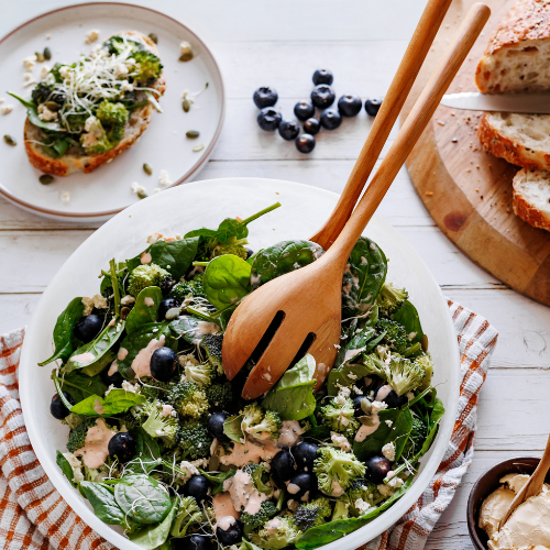 An overhead lifestyle photo of large teak wood salad servers in a white bowl of broccoli, spinach, and blueberry salad. The scene is set on a white wooden table with a side plate of avocado toast, loose blueberries, and sliced sourdough bread, showcasing a healthy, sustainable dining experience.