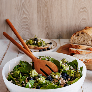 A lifestyle shot of large teak wood salad servers resting in a white ceramic bowl filled with a fresh broccoli, spinach, and blueberry salad. In the background, sliced sourdough bread sits on a wooden board, creating a healthy, rustic dining scene.
