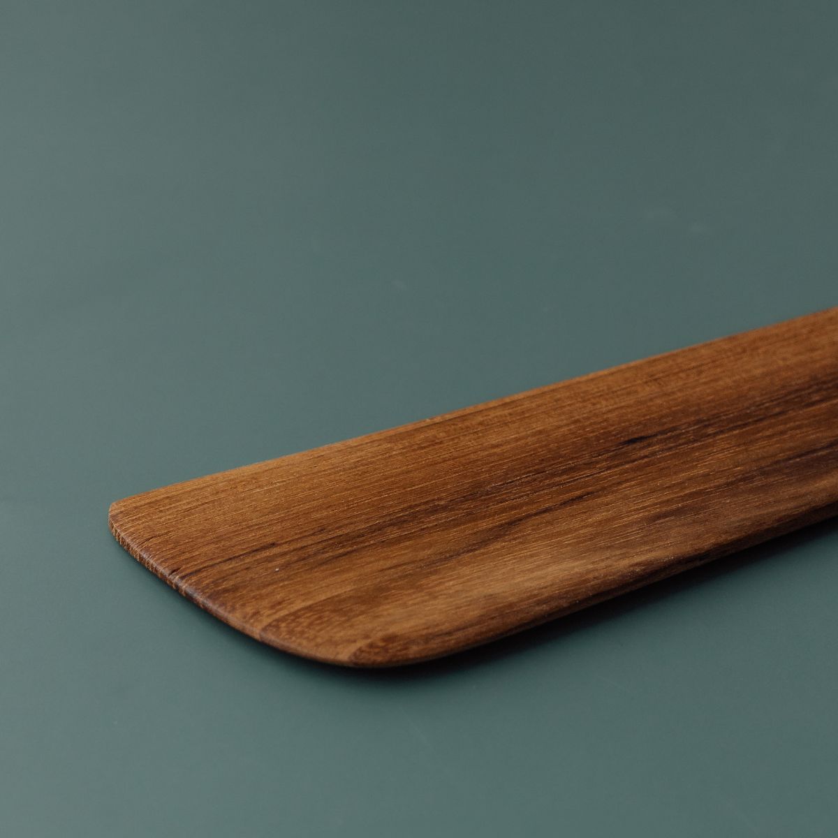 A close-up of a wooden spatula or paddle with a smooth, rounded corner and visible wood grain, resting on a solid dark teal background.