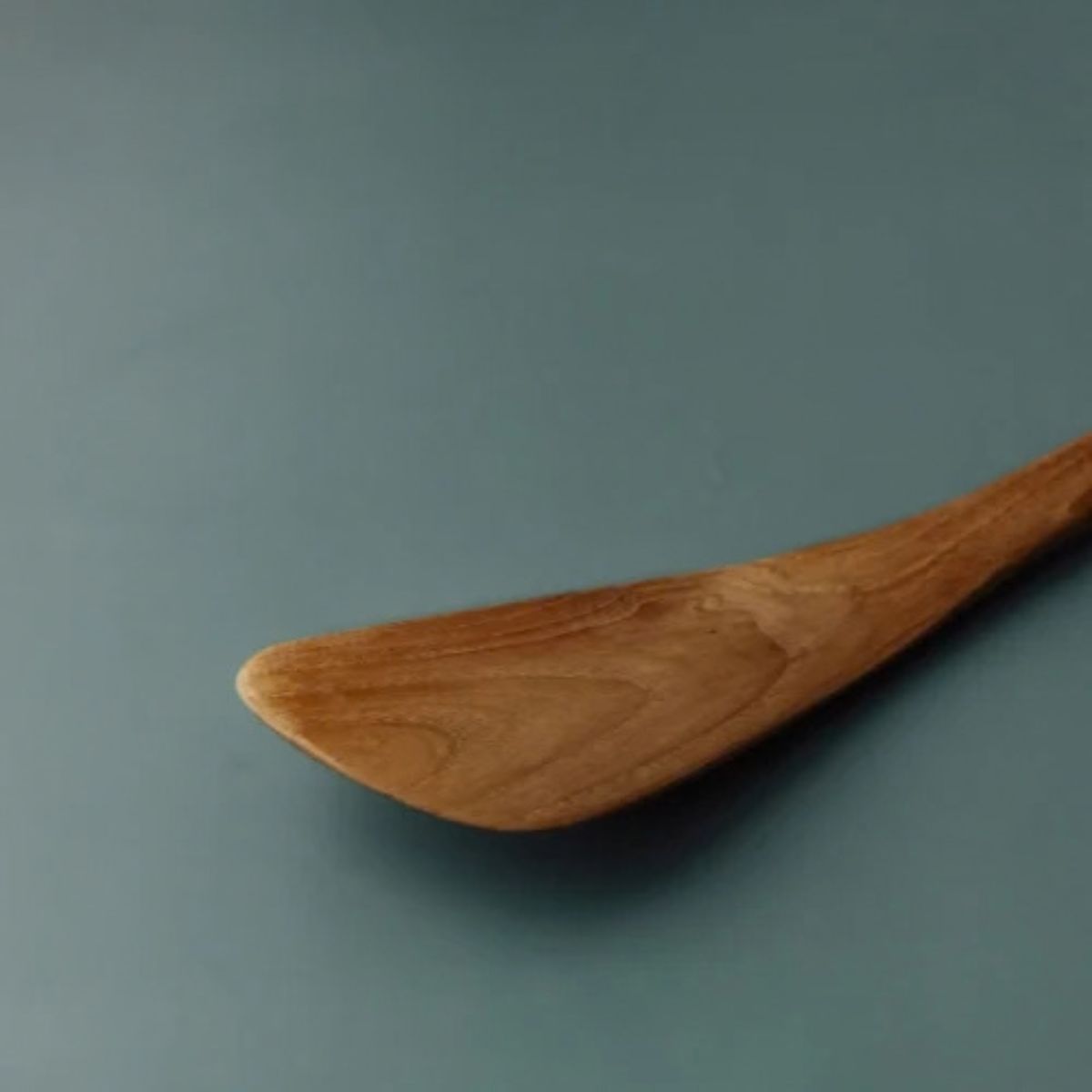A close-up of a wooden spoon or spatula with a warm brown finish and visible wood grain, resting on a smooth, muted blue-gray surface.