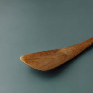 A close-up of a wooden spoon or spatula with a warm brown finish and visible wood grain, resting on a smooth, muted blue-gray surface.