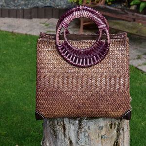 A handwoven dark brown Krajood (sea grass) bag with circular purple-tinted cane handles, displayed on a weathered tree stump in a lush green garden setting.