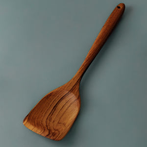 A full-length view highlighting the solid teak construction, wide paddle head, and comfortable contoured handle of our heavy-duty wooden spatula. Hand-carved from dense timber, it features a practical hanging hole for easy kitchen storage and a broad, sturdy edge designed to handle heavy frying and stirring without scratching cookware