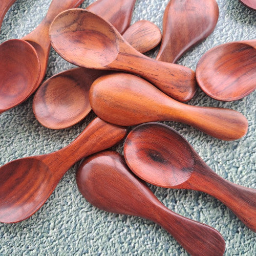 A collection of handcrafted mini rosewood spoons scattered on a textured grey surface, showcasing the vibrant reddish-brown hues and unique organic grain patterns of the solid rosewood.