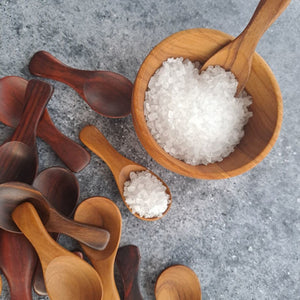 A top-down view of several handcrafted teak wood salt spoons in light and dark wood grains, with one spoon resting in a small 7 cm wooden bowl filled with coarse white sea salt on a grey textured surface.