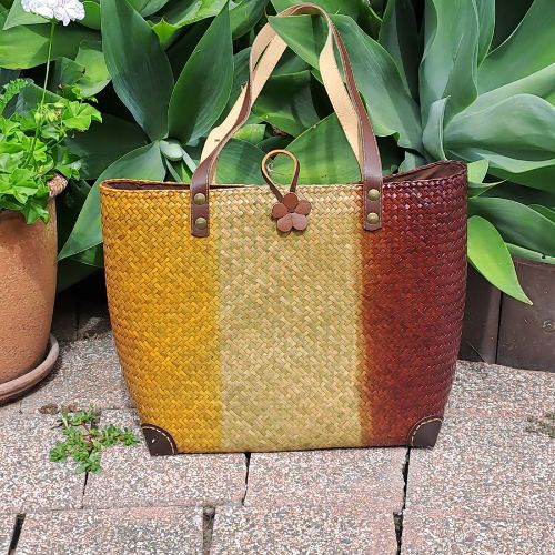 A handcrafted three-tone Krajood sea grass bag displayed outdoors on a stone patio next to a garden pot, highlighting its natural texture and earthy colors in soft daylight.