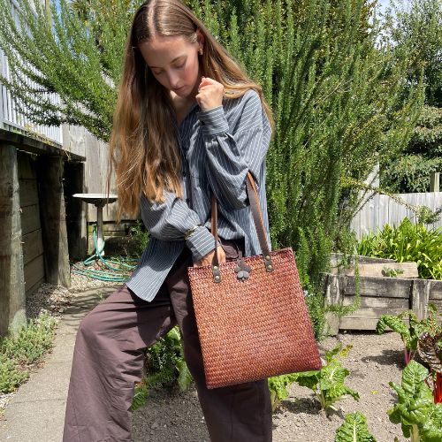 A person reaching into a mahogany brown handcrafted Krajood sea grass shoulder bag, demonstrating its spacious interior and ease of use in a garden setting.