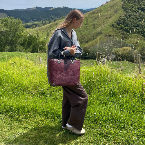 A woman walking through a grassy field, carrying a deep burgundy handcrafted Krajood sea grass tote bag with black leather handles, illustrating its durable construction and convenient size.