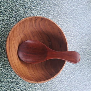A top-down view of a handcrafted dark mahogany Rosewood mini spice spoon resting inside a small, hand-turned natural teak wood pinch bowl, displaying a smooth organic wood grain contrast.