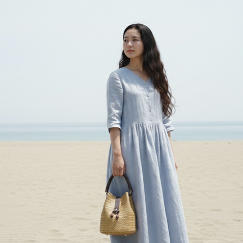 A woman in a light blue linen dress standing on a sandy beach, holding a small hand-woven Krajood bucket bag with a dark leather handle and wooden bead accents.