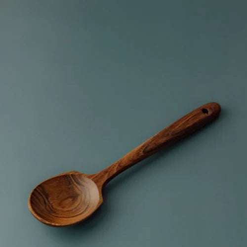 A professional product shot of a durable, hand-carved teak spoon with a wide-profile head, showcasing natural wood grain variations and a small hanging hole at the end of the tapered handle.