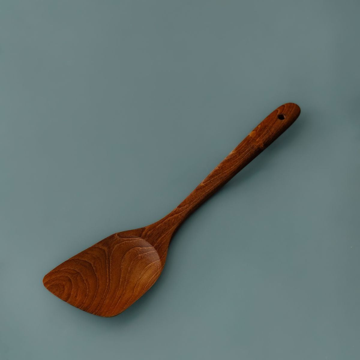 Full view of a handcrafted teak angled wooden spatula showing the complete handle with hanging hole and contoured blade against a slate blue background.