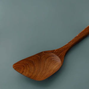 Close-up photo highlighting the natural wood grain and angled edge of a teak wooden cooking spatula against a slate blue background.