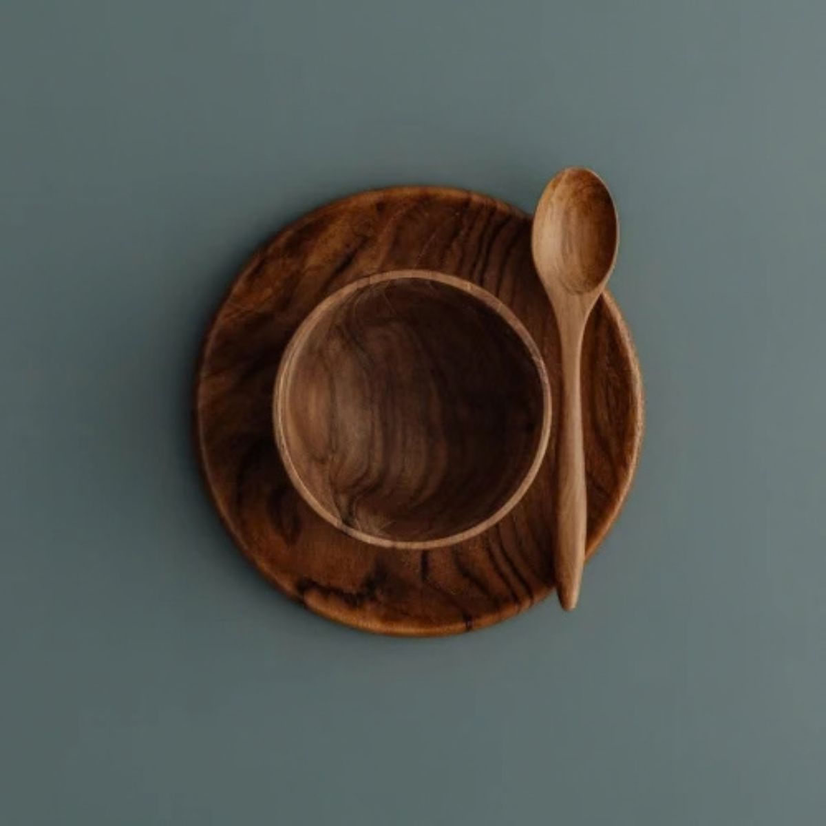 An overhead photograph of a three-piece handcrafted teak wood mealtime set for babies and toddlers, arranged on a smooth, muted grey-blue surface. The set includes a deep, rounded bowl nested atop a matching flat circular plate, with a small wooden teaspoon resting at an angle on the right. Carved from single pieces of sustainable, food-safe teak, each item displays a rich, swirling natural grain. This non-toxic and durable tableware set is designed to provide a safe, plastic-free dining experience.