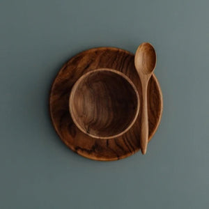 An overhead photograph of a three-piece handcrafted teak wood mealtime set for babies and toddlers, arranged on a smooth, muted grey-blue surface. The set includes a deep, rounded bowl nested atop a matching flat circular plate, with a small wooden teaspoon resting at an angle on the right. Carved from single pieces of sustainable, food-safe teak, each item displays a rich, swirling natural grain. This non-toxic and durable tableware set is designed to provide a safe, plastic-free dining experience.
