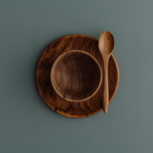 An overhead photograph of a three-piece handcrafted teak wood mealtime set for babies and toddlers, arranged on a smooth, muted grey-blue surface. The set includes a deep, rounded bowl nested atop a matching flat circular plate, with a small wooden teaspoon resting at an angle on the right. Carved from single pieces of sustainable, food-safe teak, each item displays a rich, swirling natural grain. This non-toxic and durable tableware set is designed to provide a safe, plastic-free dining experience for earl