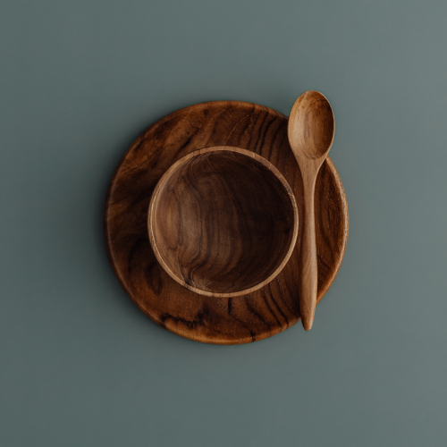 An overhead photograph of a three-piece handcrafted teak wood mealtime set for babies and toddlers, arranged on a smooth, muted grey-blue surface. The set includes a deep, rounded bowl nested atop a matching flat circular plate, with a small wooden teaspoon resting at an angle on the right. Carved from single pieces of sustainable, food-safe teak, each item displays a rich, swirling natural grain. This non-toxic and durable tableware set is designed to provide a safe, plastic-free dining experience for earl