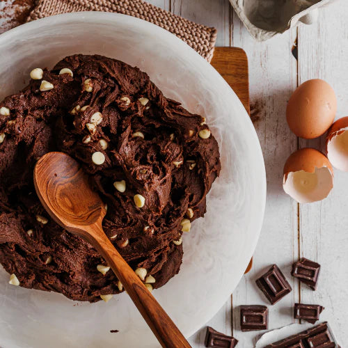 A handcrafted wide-headed teak wood spoon mixing dark chocolate cookie dough with white chocolate chips in a large white bowl, surrounded by fresh eggs and chocolate squares on a rustic white wooden surface.
