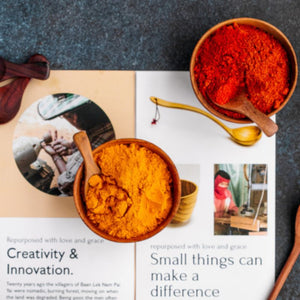 An overhead shot of two handcrafted 8 cm mini teak wood bowls filled with vibrant orange turmeric and deep red chili powder. Each bowl includes a small artisanal wooden spoon and is placed on an open book or pamphlet featuring photos of village craftspeople at work. The text "Small things can make a difference" is visible, highlighting the brand's mission of supporting artisan communities through sustainable, non-toxic kitchenware.