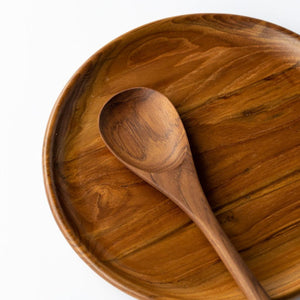 A close-up photograph of a handcrafted long-handled teak wood spoon resting on a matching large teak wood platter, presented against a clean white background. Both items showcase a deep, polished golden-brown grain with natural swirling patterns unique to high-quality teak. Carved from a single piece of sustainable, food-safe wood, this duo represents a non-toxic and durable choice for serving and entertaining in an eco-friendly home.
