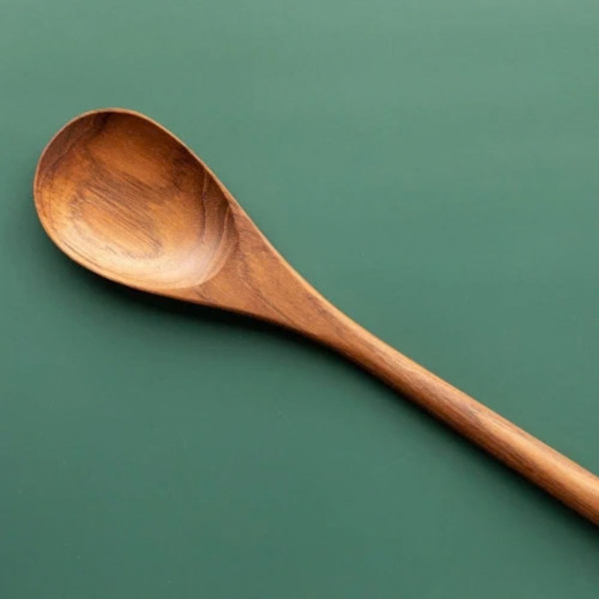 An angled overhead photograph of a single handcrafted long-handled teak wood cooking spoon resting on a smooth, muted green surface. The image focuses on the warm, rich golden-brown grain of the polished teak wood, emphasizing its smooth and food-safe finish. The shallow, rounded bowl and slender handle of this non-toxic utensil are clearly visible, showcasing it as a durable and sustainable essential for stirring pots or serving in an artisanal kitchen.