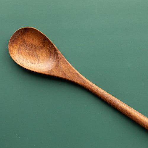 An angled overhead photograph of a single handcrafted long-handled teak wood cooking spoon resting on a smooth, muted green surface. The image focuses on the warm, rich golden-brown grain of the polished teak wood, emphasizing its smooth and food-safe finish. The shallow, rounded bowl and slender handle of this non-toxic utensil are clearly visible, showcasing it as a durable and sustainable essential for stirring pots or serving in an artisanal kitchen.