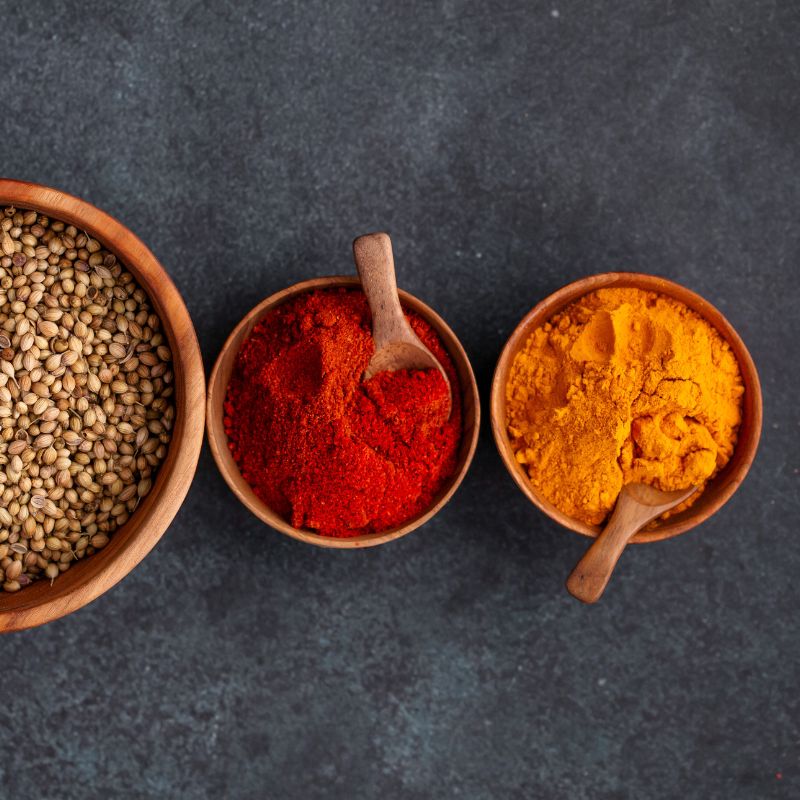 An overhead flat-lay photograph featuring three handcrafted teak wood bowls of varying sizes on a dark grey stone surface. The two smaller 8 cm mini bowls are filled with bright orange turmeric and deep red chili powder, each containing a small artisanal wooden spoon. A larger teak bowl to the left is filled with whole coriander seeds, demonstrating the rich, natural grain of the sustainably sourced wood. This clean and structured composition highlights the bowls as functional, non-toxic, and aesthetic esse