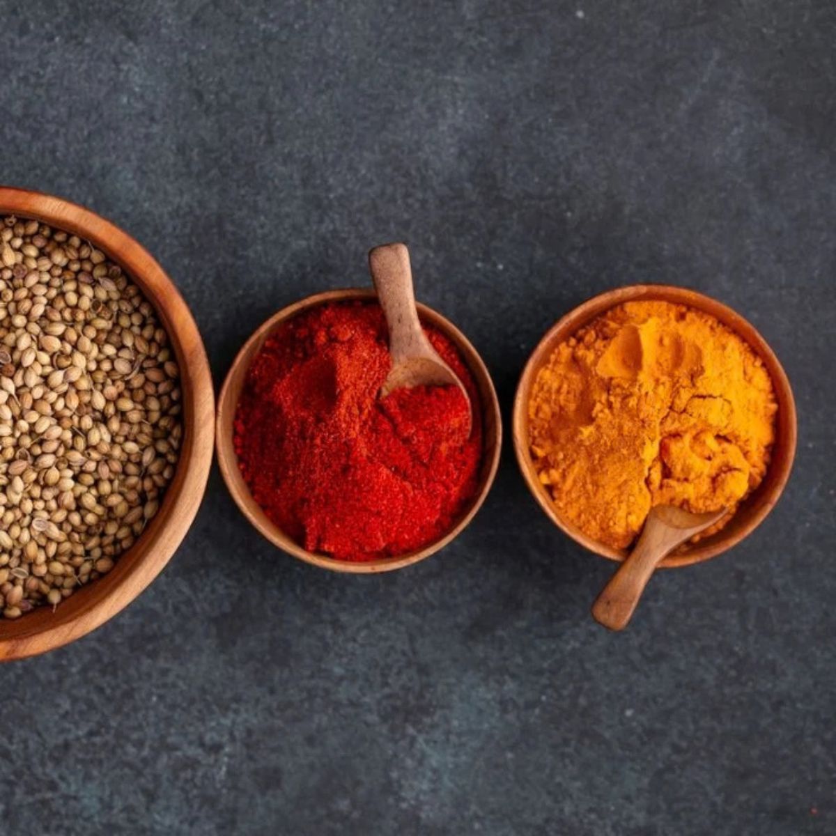 An overhead flat-lay photograph featuring three handcrafted teak wood bowls of varying sizes on a dark grey stone surface. The two smaller 8 cm mini bowls are filled with bright orange turmeric and deep red chili powder, each containing a small artisanal wooden spoon. A larger teak bowl to the left is filled with whole coriander seeds, demonstrating the rich, natural grain of the sustainably sourced wood. This clean and structured composition highlights the bowls as functional and non-toxic