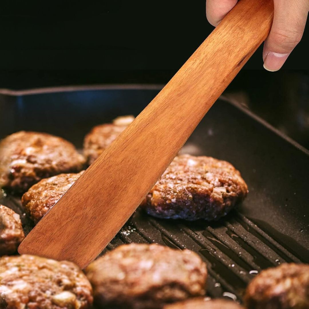 A handcrafted teak wood sauté spatula being used to flip burgers in a sizzling cast iron grill pan. The image highlights the flat-edged blade's ability to easily slide under food and the ergonomic handle designed for comfortable high-heat cooking. The rich, golden-brown teak grain contrasts beautifully with the cooking food, showcasing the spatula as both a functional and artisanal kitchen tool.