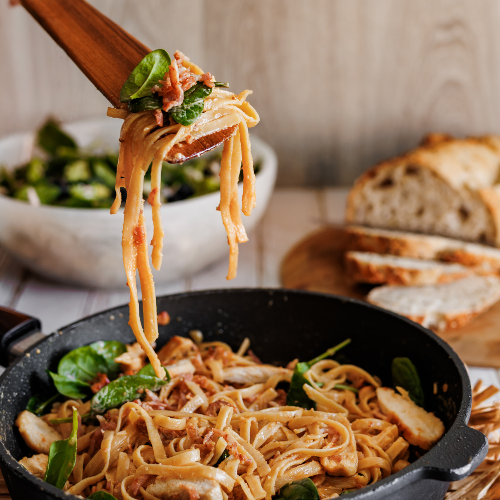 A close-up action photograph of a handcrafted teak wood sauté spatula lifting a swirl of creamy pasta, spinach, and bacon from a pan. The image focuses on the thin, tapered edge of the wooden blade as it holds the weight of the food, highlighting its strength and versatility. The warm, polished grain of the teak wood is clearly visible against the rustic home-cooked meal, emphasizing its appeal as a high-quality, sustainable kitchen tool for serious home cooks.