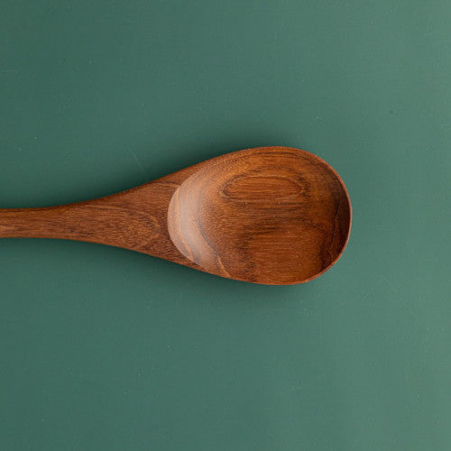 A detailed close-up photograph focusing on the head of a handcrafted teak wood spoon resting on a smooth, forest green surface. The image highlights the shallow, rounded bowl and the rich, swirling natural wood grain in warm chestnut and golden tones. The polished, food-safe finish is clearly visible, showcasing the durable and non-toxic craftsmanship of this sustainable kitchen utensil.