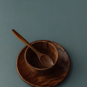 A close-up side-angle photograph of the Safer Mealtime Set, showing a small handcrafted teak wood teaspoon resting at an upward angle inside a deep, rounded 11 cm bowl. The bowl is nested on a matching flat teak plate, all set against a smooth, muted grey-blue background. The image highlights the ergonomic handle of the spoon and the rich, swirling natural grain of the sustainable, food-safe wood. This non-toxic and durable tableware set is designed to support independent eating for babies and toddlers.
