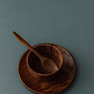 A close-up side-angle photograph of the Safer Mealtime Set, showing a small handcrafted teak wood teaspoon resting at an upward angle inside a deep, rounded 11 cm bowl. The bowl is nested on a matching flat teak plate, all set against a smooth, muted grey-blue background. The image highlights the ergonomic handle of the spoon and the rich, swirling natural grain of the sustainable, food-safe wood. This non-toxic and durable tableware set is designed to support independent eating for babies and toddlers.