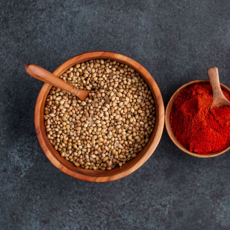 An overhead photograph of two handcrafted teak wood bowls of differing sizes resting on a dark, slate-grey surface. The larger 13 cm bowl is filled with whole coriander seeds and contains a small wooden spoon, highlighting its deep, rounded basin. The smaller bowl to the right is filled with bright red paprika. The rich, warm natural grain of the sustainable teak wood is visible on both pieces, showcasing them as non-toxic, food-safe, and durable essentials for spice storage or serving in an artisanal kitch