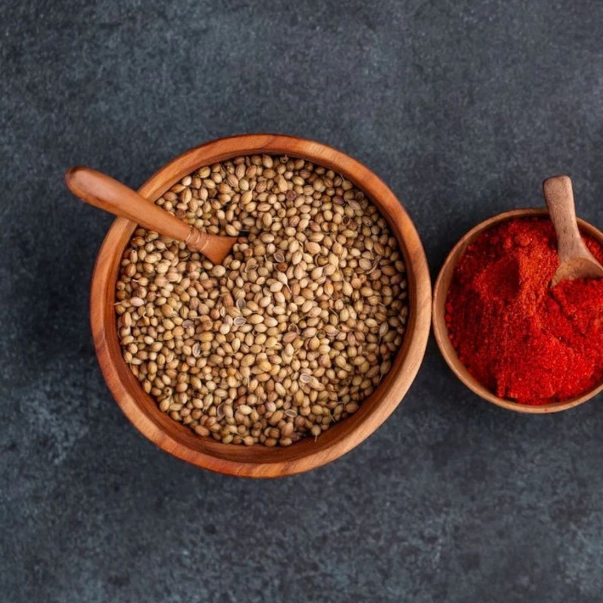 An overhead photograph of two handcrafted teak wood bowls of differing sizes resting on a dark, slate-grey surface. The larger 13 cm bowl is filled with whole coriander seeds and contains a small wooden spoon, highlighting its deep, rounded basin. The smaller bowl to the right is filled with bright red paprika. The rich, warm natural grain of the sustainable teak wood is visible on both pieces, showcasing them as non-toxic, food-safe, and durable essentials for spice storage.