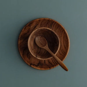 A side-angle photograph of the three-piece handcrafted teak wood Safer Mealtime Set for babies and toddlers. The deep, rounded bowl is nested on a flat circular plate, with a small artisanal teaspootic-free tableware set is designed for durability and safety in early childhood dining.n resting beside them on a muted grey-blue surface. The perspective highlights the solid, single-piece construction and the rich, natural wood grain of the sustainable teak. This non-toxic