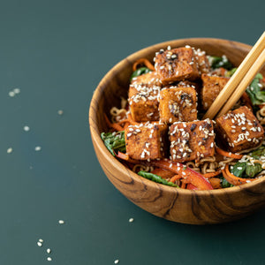 A close-up lifestyle photograph of a 13 cm handcrafted teak wood bowl filled with a colorful tofu and noodle stir-fry, topped with sesame seeds. A pair of wooden chopsticks rests in the bowl, highlighting its depth and rounded interior. The rich, swirling natural grain of the sustainable teak wood is visible against a deep forest green background, showcasing the bowl as a durable, non-toxic, and aesthetic choice for daily meals.