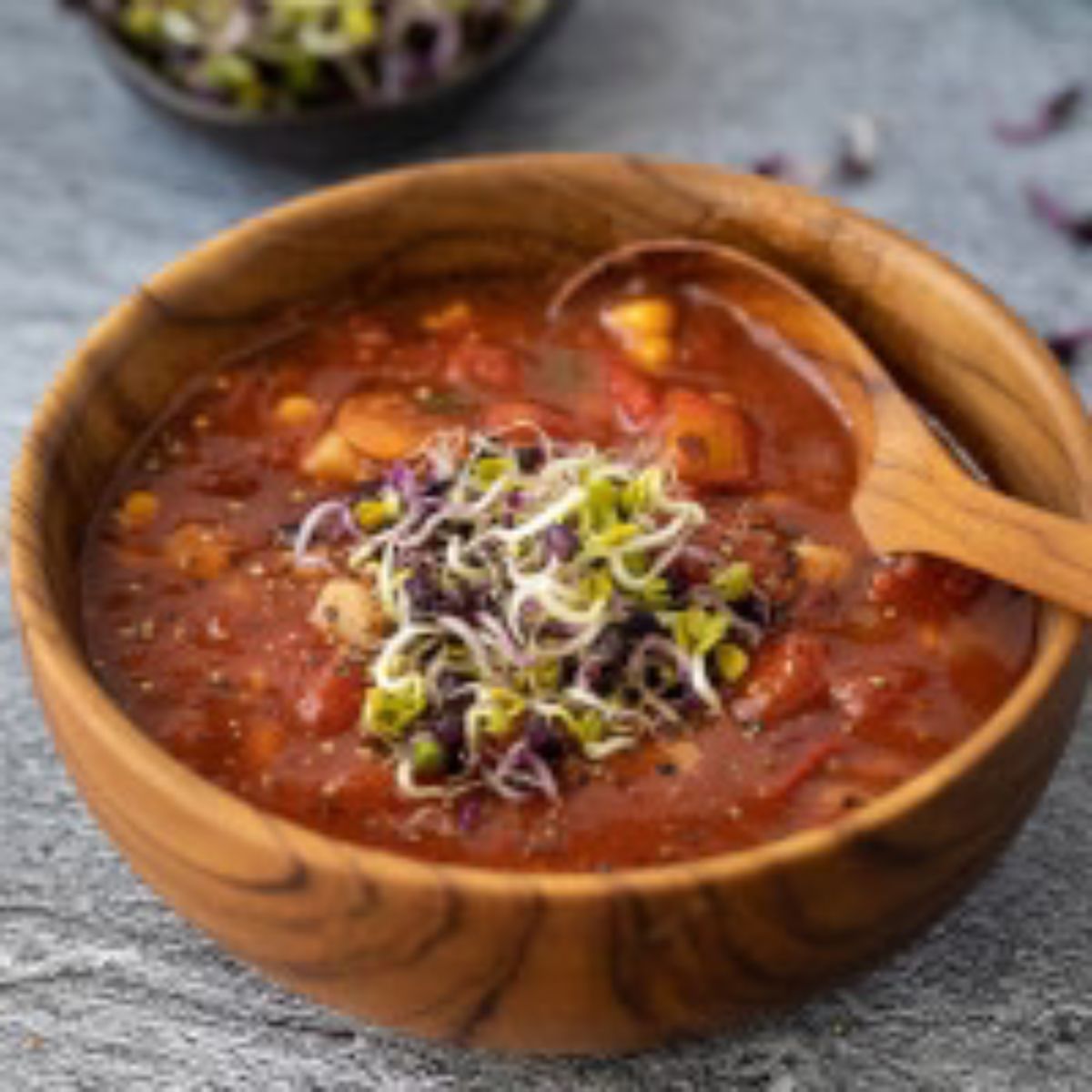 A close-up lifestyle photograph focusing on a handcrafted 13 cm teak wood bowl filled with a hearty, chunky red vegetable and bean stew. The stew is topped with fresh green and purple sprouts and includes corn kernels and black beans. A large, matching teak wood spoon rests inside the bowl, highlighting its generous depth and smooth, rounded interior basin. The rich, natural grain of the sustainable teak wood is prominent on both the bowl and spoon, set against a muted grey stone surface.