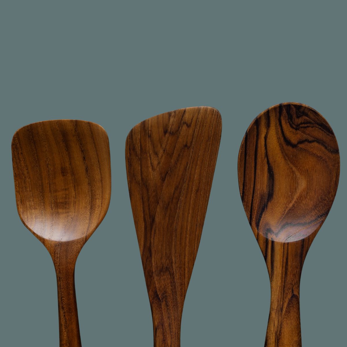 A close-up top-down product photograph displaying the working heads of three handcrafted teak wood cooking utensils lined up side-by-side against a smooth, solid slate-grey background. From left to right: a wide rectangular cooking spatula, a flat asymmetric sauté scraper spatula, and a deep oval cooking spoon. The image showcases the varied golden-brown hues and striking dark, swirling natural wood grain patterns of each single-piece, sustainable wooden tool.