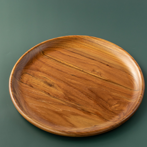 A top-down product photograph of a handcrafted round dining plate carved from sustainable teak wood, displayed against a smooth, dark forest-green background. The image clearly showcases the warm, golden-brown timber tones and prominent, swirling natural wood grain. The plate features a smooth, slightly raised rim and a food-safe, non-toxic finish, highlighting its intentional and durable design for daily plastic-free meals.
