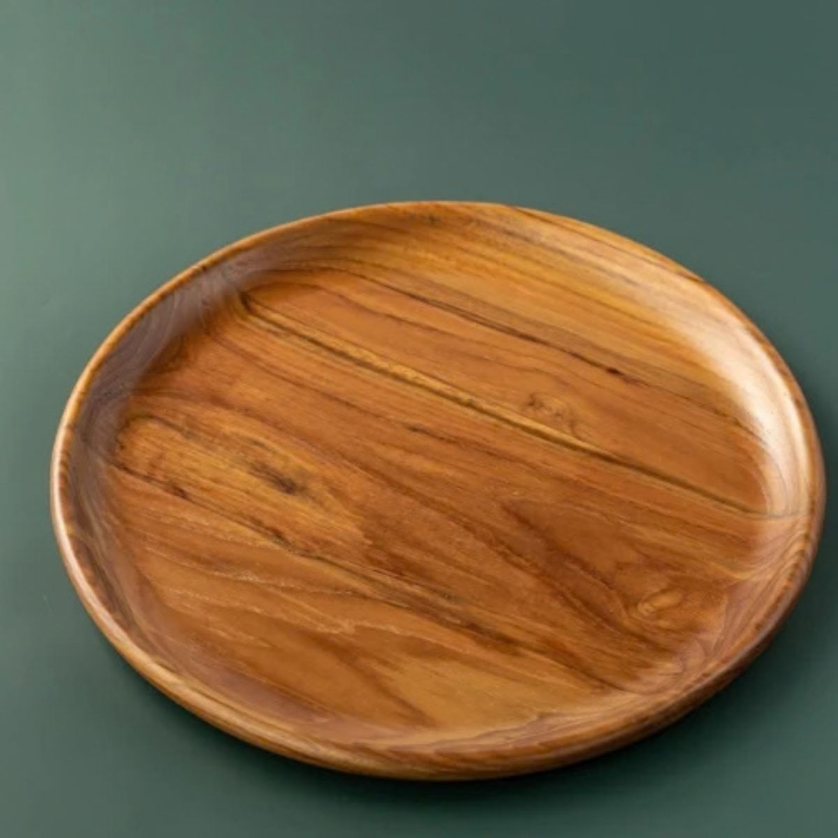 A top-down product photograph of a handcrafted round dining plate carved from sustainable teak wood, displayed against a smooth, dark forest-green background. The image clearly showcases the warm, golden-brown timber tones and prominent, swirling natural wood grain. The plate features a smooth, slightly raised rim and a food-safe, non-toxic finish, highlighting its intentional and durable design for daily plastic-free meals.