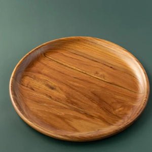 A top-down product photograph of a handcrafted round dining plate carved from sustainable teak wood, displayed against a smooth, dark forest-green background. The image clearly showcases the warm, golden-brown timber tones and prominent, swirling natural wood grain. The plate features a smooth, slightly raised rim and a food-safe, non-toxic finish, highlighting its intentional and durable design for daily plastic-free meals.