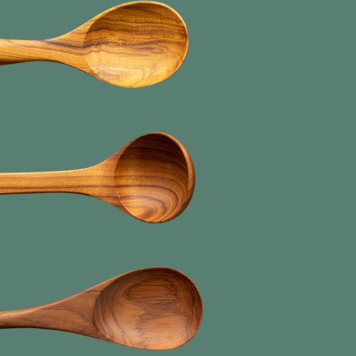 A top-down product photograph showcasing a trio of handcrafted teak wood serving utensils aligned horizontally along the left side against a smooth, solid sage-green background. The image displays the bowls of three distinct utensils—a wide shallow spoon, a rounded deep ladle spoon, and a large tapered serving spoon—each carved from sustainable timber. The composition highlights the varied golden-brown tones and rich, flowing natural wood grain patterns inherent to each single-piece, non-toxic kitchen tool.